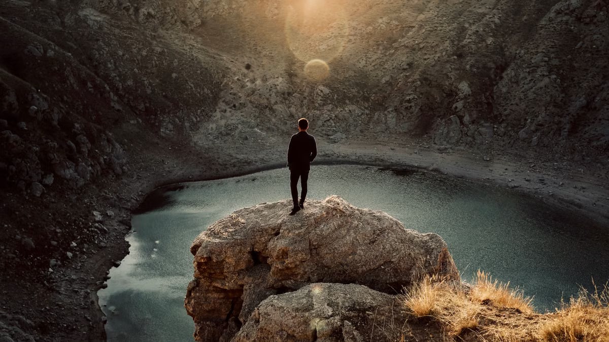 A man stands on a rocky cliff overlooking a tranquil lake in a remote canyon at sunset.